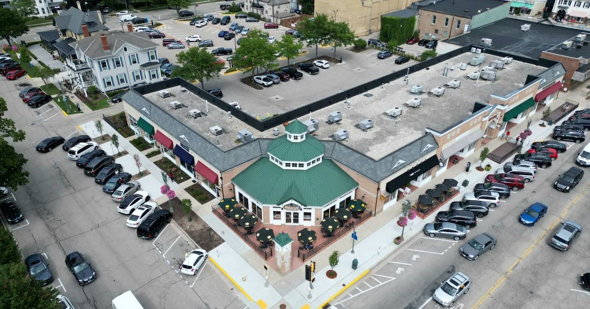 Bird's-eye view of a bustling suburban shopping center with parked cars and tree-lined streets.
